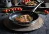 A cooked steak in a frying pan, surrounded by ingredients like tomatoes, jalapeños, and peppercorns. Produktbild-Vorschau