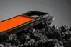 A close-up shot of an orange and black phone case resting on dark, textured rocks. Podgląd zdjęcia produktu