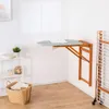 A wall-mounted ironing board is shown in a laundry room setting, with a drying rack and a basket of laundry nearby. Voir les images du produit