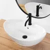 A white oval countertop sink with a black faucet is shown on a wooden surface, with a reflection of the sink and faucet visible in a round mirror. Náhled obrázku produktu