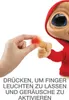 A hand is shown pressing the finger of a plush toy E.T., which is glowing red. The toy is wearing a red hooded jacket. Produktbild-Vorschau