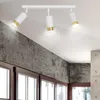A white ceiling track light with three adjustable spotlights, each with a white cylindrical shade and a gold accent ring, is mounted on a white ceiling. The background includes a textured gray wall and wooden window frames. Podgląd zdjęcia produktu