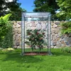 A small greenhouse with tomato plants inside, set in a garden with a stone wall and grass. Náhled obrázku produktu