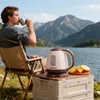 A man is drinking from a mug while sitting in a camping chair next to a lake with mountains in the background. A portable electric kettle and a plate of fruit are on a storage box in the foreground. Podgląd zdjęcia produktu