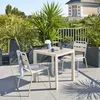 A patio set with a table and chairs on a tiled balcony, surrounded by potted plants and overlooking a building and trees. Produktbild-Vorschau