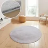 A round, taupe-silver rug is placed on a light wood floor in a room with a window, a chair, and a woven stool. Produktbild-Vorschau