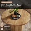 A round wooden coffee table with decorative items on top, set in a living room environment. Produktbild-Vorschau