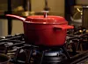 A red cast iron pot with a silver lid handle is on a gas stove with a blue flame underneath. Anteprima dell’immagine prodotto