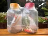 Two large glass jars with red and yellow lids, shrink-wrapped together, sitting on a wooden surface outdoors with plants in the background. Produktbild-Vorschau