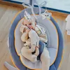 A baby is lying in a gray baby bouncer with orange trim, with toys hanging above. Produktbild-Vorschau