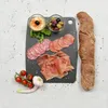 A charcuterie board with various meats, olives, hummus, tomatoes, garlic, and a baguette on a black cutting board. Náhľad obrázku produktu