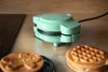 A mint green waffle maker is shown on a wooden countertop next to two freshly made waffles. Part of a black stovetop is visible to the left. Podgląd zdjęcia produktu