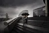 A couple walks down stairs under a black umbrella on a cloudy day, with iconic London landmarks in the background. Anteprima dell’immagine prodotto