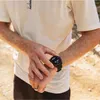 A person wearing a white t-shirt and brown pants checks their black smartwatch. Náhľad obrázku produktu