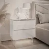 A white, high-gloss nightstand with chrome legs, decorated with a bust and a lamp, next to a bed. Produktbild-Vorschau