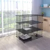 A metal cage for pets is set up in a room with a large window showing a garden. The cage has multiple levels and includes a pet bed and a food bowl. Podgląd zdjęcia produktu