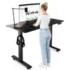A woman is standing at a black adjustable height desk with a computer monitor, keyboard, and lamp. Headphones are hanging from the desk. Produktbild-Vorschau