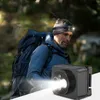 A man wearing a headlamp and backpack in a forest setting, with a small black device in the foreground emitting a bright light. Náhled obrázku produktu