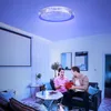 A couple is sitting on a couch in a living room, with one person handing a gift to the other. The room is lit by a modern ceiling light with a blue hue. Produktbild-Vorschau