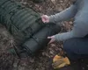A person is adjusting straps on a large green backpack in a forest setting. Produktbild-Vorschau
