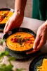 Hands holding a bowl of soup with carrots and pine nuts. Náhľad obrázku produktu