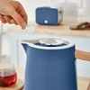 A hand is adding something to a blue kettle, with a toaster and a cup of tea in the background. Produktbild-Vorschau