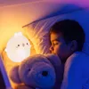 A young boy sleeps in bed next to a glowing bunny-shaped night light and a teddy bear. Produktbild-Vorschau
