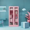 A pink metal cabinet with glass doors is displayed in a studio setting with a blue wall and floor. Produktbild-Vorschau