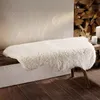 A fluffy beige sheepskin rug is draped over a wooden bench in a rustic setting with a pile of firewood in the background and candles on a shelf. Produktbild-Vorschau