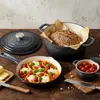 A collection of cast iron cookware, including a Dutch oven with bread, a frying pan with a vegetable dish, and a small ramekin with tomatoes, arranged on a wooden table. Produktbild-Vorschau
