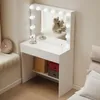 A white vanity table with LED lights and a mirror, set up in a room with a window seat and a bed. It has makeup items on top and storage below. Podgląd zdjęcia produktu