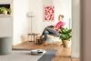 A woman is reading a book while a robot vacuum cleaner is cleaning the floor in a living room. Produktbild-Vorschau