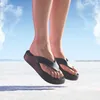 A person's feet wearing black platform flip-flops are shown jumping in the air over a white, flat surface with a blue sky and clouds in the background. Voir les images du produit