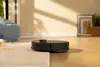 A black robot vacuum cleaner is cleaning a floor in a room with natural light casting shadows. Produktbild-Vorschau