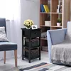 A black nightstand with a drawer and two baskets is placed next to a blue chair and a bookshelf in a room. Náhled obrázku produktu