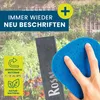 A hand is holding a blue sponge to a dark slate plant marker with text on it, set against a garden background. Produktbild-Vorschau