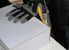 A person wearing black gloves is using a utility knife to open a white cardboard box. Produktbild-Vorschau