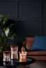 Three Woodwick candles are displayed on a dark table in front of a dark blue paneled wall and a brown leather sofa with a blue pillow. Produktbild-Vorschau