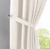 A close-up of a cream-colored curtain held back by a fabric tie and a metal hook, with a blurred green tree visible through the window. Produktbild-Vorschau