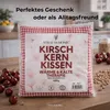 A packaged cherry stone pillow with cherries and loose cherry stones in the foreground and background. Produktbild-Vorschau
