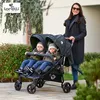 A woman pushing a twin stroller with two babies in it, outdoors. Produktbild-Vorschau