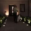 A woman walks up a illuminated pathway towards a house at night, carrying shopping bags. Produktbild-Vorschau