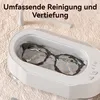 A white ultrasonic cleaner with glasses inside, next to two toothbrushes on a textured fabric background. Produktbild-Vorschau