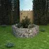 A hexagonal gabion raised garden bed filled with plants, set in a grassy backyard with a wooden fence in the background. Podgląd zdjęcia produktu