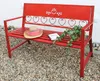 A red metal garden bench with decorative heart cutouts on the backrest, placed on a gravel surface next to a white wall. A straw hat with a pink scarf and a small book are on the seat, along with a potted plant. Produktbild-Vorschau