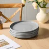 A stack of four gray plates on a wooden table, with a chair and flowers in the background. Produktbild-Vorschau