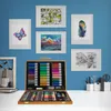 An art supply set in a wooden case is displayed on a white desk against a blue wall with framed artwork. Náhled obrázku produktu