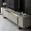 A modern TV stand with a travertine-like finish and gold accents, placed in a dimly lit room. Voir les images du produit