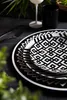 A close-up shot of a stack of black and white patterned plates on a dark tablecloth, with greenery in the foreground and background. Produktbild-Vorschau