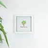 A white framed picture with a tree illustration and text is displayed on a white wall, with a blurred green plant in the foreground. Produktbild-Vorschau
